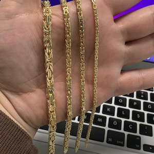May include: Four gold-colored Byzantine chain necklaces of varying widths are displayed. The chains are held in a hand, showcasing their intricate link design and polished finish. The necklaces are a warm, yellow gold color.