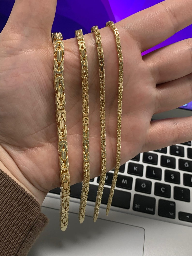 May include: Four gold-colored Byzantine chain necklaces of varying widths are displayed. The chains are held in a hand, showcasing their intricate link design and polished finish. The necklaces are a warm, yellow gold color.