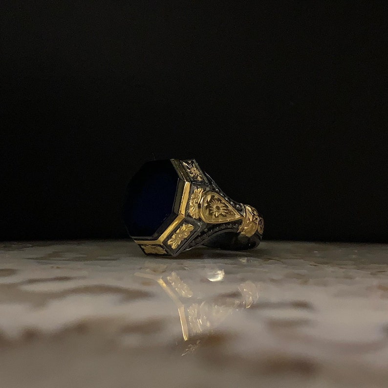 May include: A detailed close-up of a ring with a dark blue, octagonal stone. The ring has a gold-colored filigree design on a dark metal band. The ring is set against a dark background, with a reflective surface.