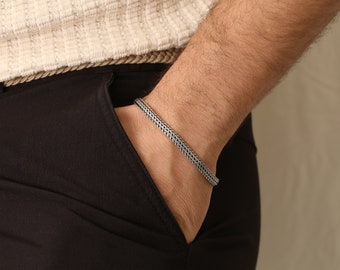 May include: A silver bracelet with a woven chain design. The bracelet is worn on a wrist, with the arm resting in a black pocket. The person is wearing a cream-colored sweater and black pants. The bracelet is a simple, yet elegant piece of jewelry.