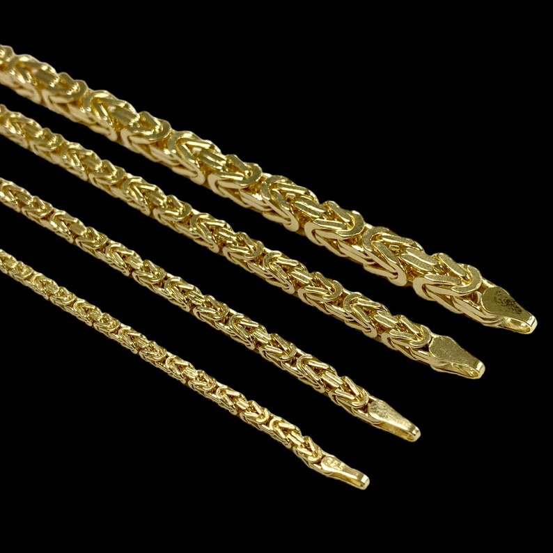 May include: Four gold-colored Byzantine chain necklaces of varying thicknesses are displayed against a black background. The chains are made of interlocking links, creating a textured surface. The chains are angled diagonally across the frame.