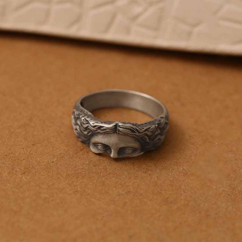 K&ouml;nnte beinhalten: Ein silberfarbener Metallring mit einem skulptierten menschlichen Gesicht und detailliertem Haar, das sich entlang des Rings erstreckt. Die Oberfl&auml;che hat ein oxidiertes Finish, das die Gesichtsz&uuml;ge und die Haarstruktur plastisch hervorhebt.