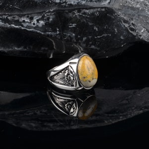 Peut inclure: Une bague en argent avec une pierre ovale jaune et blanche sertie dans une bande en argent. La bande pr&eacute;sente un motif d&eacute;taill&eacute; d'ailes de chaque c&ocirc;t&eacute; de la pierre.