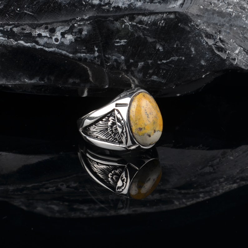 Peut inclure: Une bague en argent avec une pierre ovale jaune et blanche sertie dans une bande en argent. La bande pr&eacute;sente un motif d&eacute;taill&eacute; d'ailes de chaque c&ocirc;t&eacute; de la pierre.