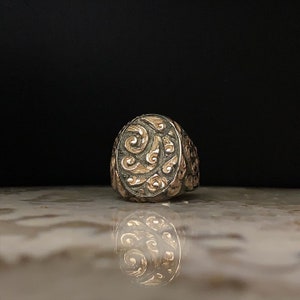 May include: A detailed close-up of a silver and gold ring. The ring features an oval face with intricate swirling patterns in gold against a silver background. The ring's design is symmetrical and ornate, reflecting light.