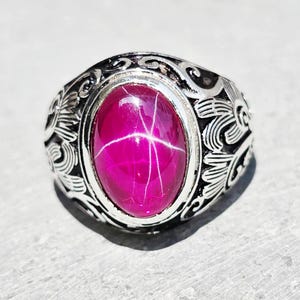May include: A silver ring featuring an oval, magenta gemstone with a star effect. The ring's band has an ornate, swirling pattern. The gemstone is set in a silver bezel. The ring is displayed on a light grey surface.
