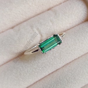 May include: A silver ring with a green emerald-cut gemstone. The ring is displayed in a white ring box.