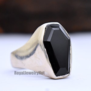 May include: A silver ring with a black coffin-shaped gemstone. The ring is on a white surface.