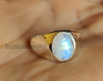 Puede incluir: Un anillo de plata con una piedra lunar ovalada e iridiscente. La piedra lunar muestra un juego de colores azules y blancos. El anillo se muestra en una mano, enfocando la joya.