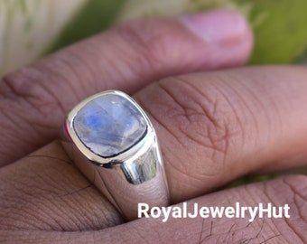Puede incluir: Un anillo de plata con una piedra de luna de forma cuadrada. El anillo tiene una banda gruesa y un acabado liso y pulido.