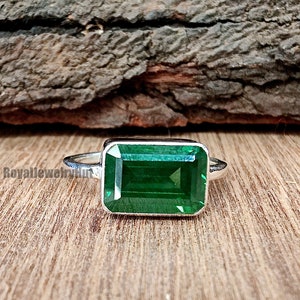 Könnte beinhalten: Ein silberner Ring mit einem großen, rechteckigen, smaragdgeschliffenen grünen Edelstein. Der Ring ist auf einem silbernen Band gefasst und wird auf einer Holzoberfläche präsentiert. Das Design des Rings unterstreicht das leuchtende Grün des Steins.
