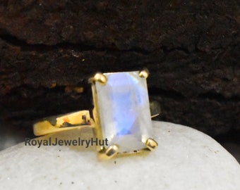 Puede incluir: Un anillo de oro con una piedra de luna rectangular e iridiscente. El anillo está colocado sobre una superficie de piedra blanca.
