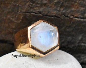 Puede incluir: Un anillo de oro con una gran piedra de luna hexagonal e iridiscente. El anillo está colocado sobre una piedra blanca.
