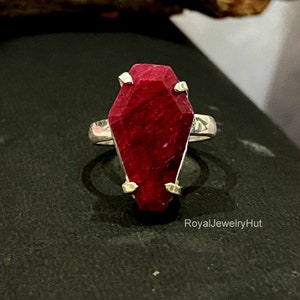 Puede incluir: Un anillo de plata con una gran piedra preciosa de rubí rojo facetado engastado en un diseño en forma de ataúd. El anillo está sobre un fondo negro. El texto 'RoyalJewelryHut' está impreso en blanco en la parte inferior de la imagen.