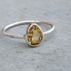 Puede incluir: Un anillo de plata con una piedra preciosa de color amarillo dorado en forma de pera. La piedra preciosa está engastada en un engaste de bisel, y la banda es fina y pulida. El anillo se muestra sobre una superficie gris.