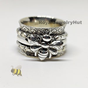 Może przedstawiać: Srebrny pierścień obrotowy z motywem pszczoły. Pierścień ma wiele pasków z misternymi wzorami.