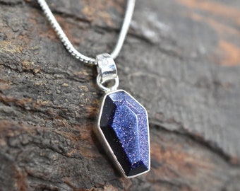 May include: A silver chain necklace with a blue gemstone pendant shaped like a coffin. The gemstone has a sparkly, glittery texture.