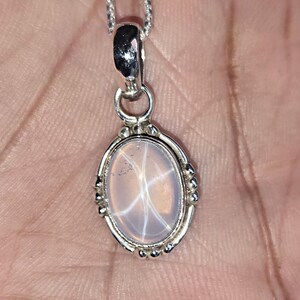 May include: A silver pendant necklace with an oval, pale gemstone displaying a star pattern. The pendant is set in a silver frame with decorative details and is suspended from a fine silver chain.