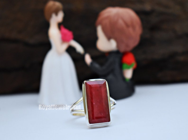 May include: A silver ring featuring a large, rectangular, red gemstone. The ring is in focus, with a pair of figurines blurred in the background.