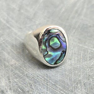 May include: A silver ring with an oval-shaped abalone shell inlay. The shell has a green, blue, and purple iridescent pattern.