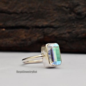 May include: A silver ring with a clear, iridescent rectangular gemstone. The ring is set on a white background.