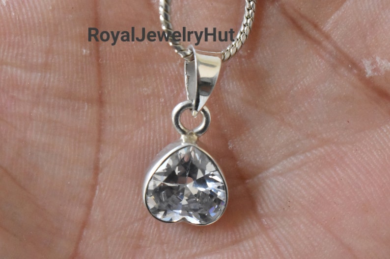 Puede incluir: Un colgante de plata en forma de coraz&oacute;n con una piedra de cristal transparente. El colgante est&aacute; unido a una cadena de plata.