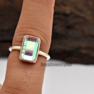 May include: A silver ring with a rectangular iridescent gemstone. The gemstone has a light blue, green, and purple hue. The ring is on a finger.