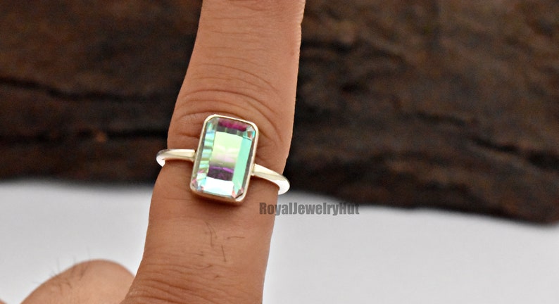 May include: A silver ring with a rectangular iridescent gemstone. The gemstone has a light blue, green, and purple hue. The ring is on a finger.