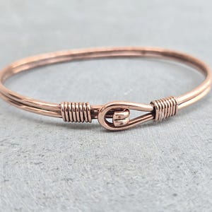 May include: A copper-coloured bangle bracelet with a hook and eye clasp. The bracelet is made of thin, round wire and features wrapped wire detailing near the clasp. The clasp is a simple, functional design. The bracelet is shown on a grey surface.