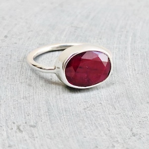 May include: A silver ring featuring a large, oval-shaped ruby gemstone. The ruby is a deep red colour and has a faceted cut.