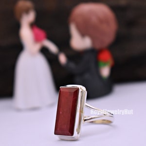 May include: A silver ring featuring a rectangular red gemstone. The ring is set against a blurred background of a bride and groom figurine.