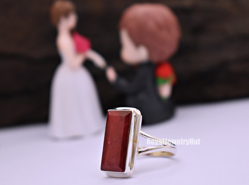May include: A silver ring featuring a rectangular red gemstone. The ring is set against a blurred background of a bride and groom figurine.