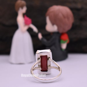 May include: A silver ring featuring a rectangular red gemstone. The ring is set against a blurred background of a bride and groom figurine.
