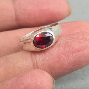Puede incluir: Un anillo de plata con una gran piedra preciosa ovalada de color rojo intenso. La piedra preciosa está engastada en una banda de plata lisa y pulida. El diseño del anillo es simple pero elegante, mostrando el color vibrante de la piedra.