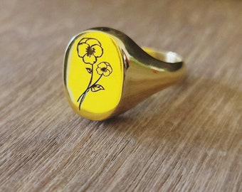 May include: A gold signet ring with an engraved design of two yellow flowers with black outlines.