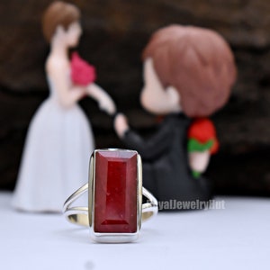 May include: A silver ring featuring a rectangular red gemstone. The ring is in focus, with a blurred image of a bride and groom figurine in the background.
