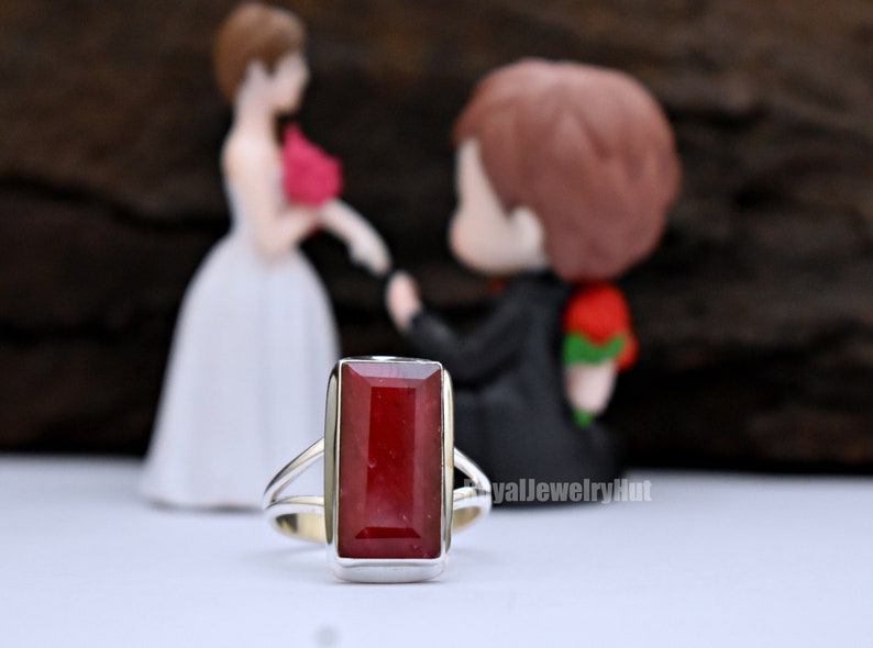 May include: A silver ring featuring a rectangular red gemstone. The ring is in focus, with a blurred image of a bride and groom figurine in the background.