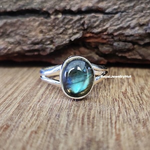 Könnte beinhalten: Ein silberner Ring mit einem ovalen Labradorit-Edelstein. Der Edelstein zeigt schillernde blaue und grüne Farbtöne. Der Ring hat ein geteiltes Schaftdesign. Der Ring befindet sich auf einer Holzoberfläche. Der Text "RoyalJewelryHut" ist sichtbar.