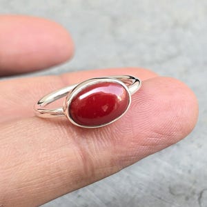 May include: A silver ring with a red oval gemstone. The ring is on a finger.