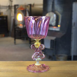 May include: A handcrafted, pink glass goblet with a tulip-shaped bowl and a decorative stem. The stem features intricate swirls of red, white, and clear glass. The goblet sits on a round, pink base. The background shows a workshop setting.