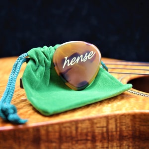 Puede incluir: Una púa de guitarra de caparazón de tortuga marrón con la palabra "hense" inscrita en letras doradas. La púa está apoyada en una bolsa de terciopelo verde sobre una superficie de madera.