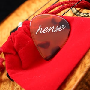 Puede incluir: Una púa de guitarra de carey con la palabra "hense" en letras blancas descansa sobre una bolsa de terciopelo rojo. La púa es triangular y tiene un acabado brillante. Se ve un cordón rojo.