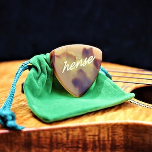 May include: A brown and white guitar pick with the word "hense" inscribed on it. The pick is sitting on a green velvet pouch on top of a wooden surface.