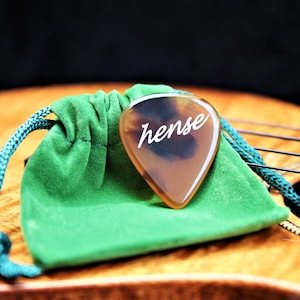 May include: A brown guitar pick with the word "hense" written on it in white. The pick is in a green velvet pouch.