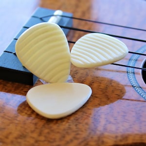 May include: Three white guitar picks with a ridged texture, lying on a wooden ukulele.
