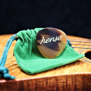 May include: A brown and black guitar pick with the word "hense" inscribed in white lettering. The pick is resting on a green velvet pouch.