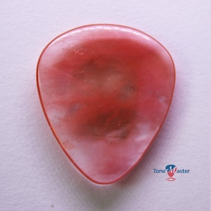 Puede incluir: Una púa de guitarra rosa con un patrón de remolino blanco. La púa está hecha por Tone Master USA.