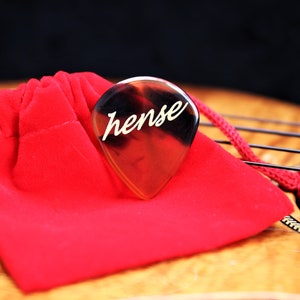 Puede incluir: Una púa de guitarra transparente marrón con la palabra "hense" inscrita en letras doradas. La púa está apoyada en una bolsa de terciopelo rojo.