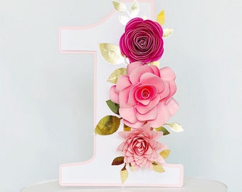 Könnte beinhalten: Weißer Kuchen mit einem rosa und goldenen Papierblumen-Topper in Form der Zahl eins. Der Kuchen steht auf einem gläsernen Kuchenständer.
