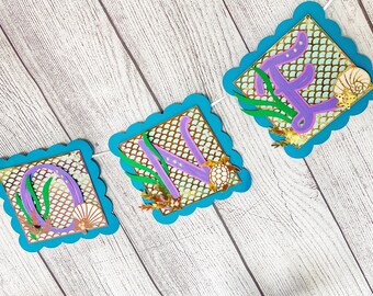 Puede incluir: Un banner "UNO" azul y morado con temática de sirena. Las letras son moradas con detalles de purpurina dorada. El fondo es un patrón de escamas de sirena de purpurina dorada. Hay algas verdes y conchas.